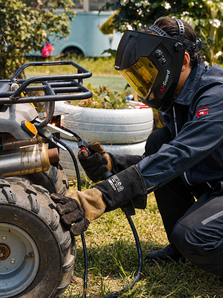 A person wearing a black welding helmet with a yellow shield, black welding jacket, and protective gloves kneels beside a vehicle tire outdoors, holding a welding torch near the tire.