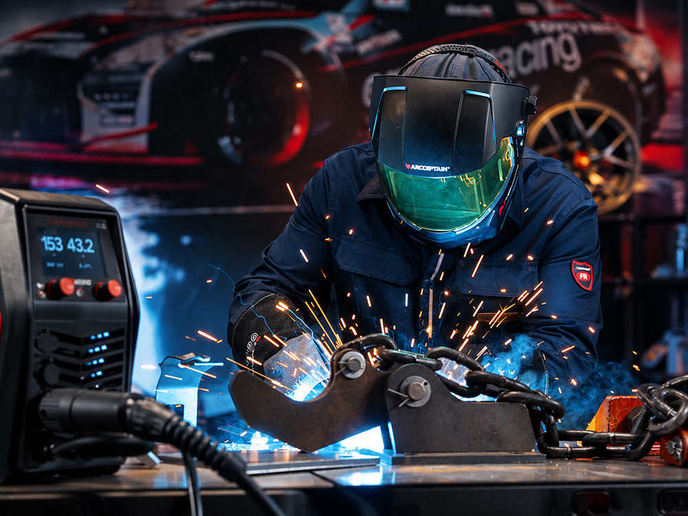 A welder in a dark blue jumpsuit and welding helmet generates bright blue light and orange sparks while working on a metal piece on a workshop table, with a welding machine visible on the left and a blurred racing car poster in the background.